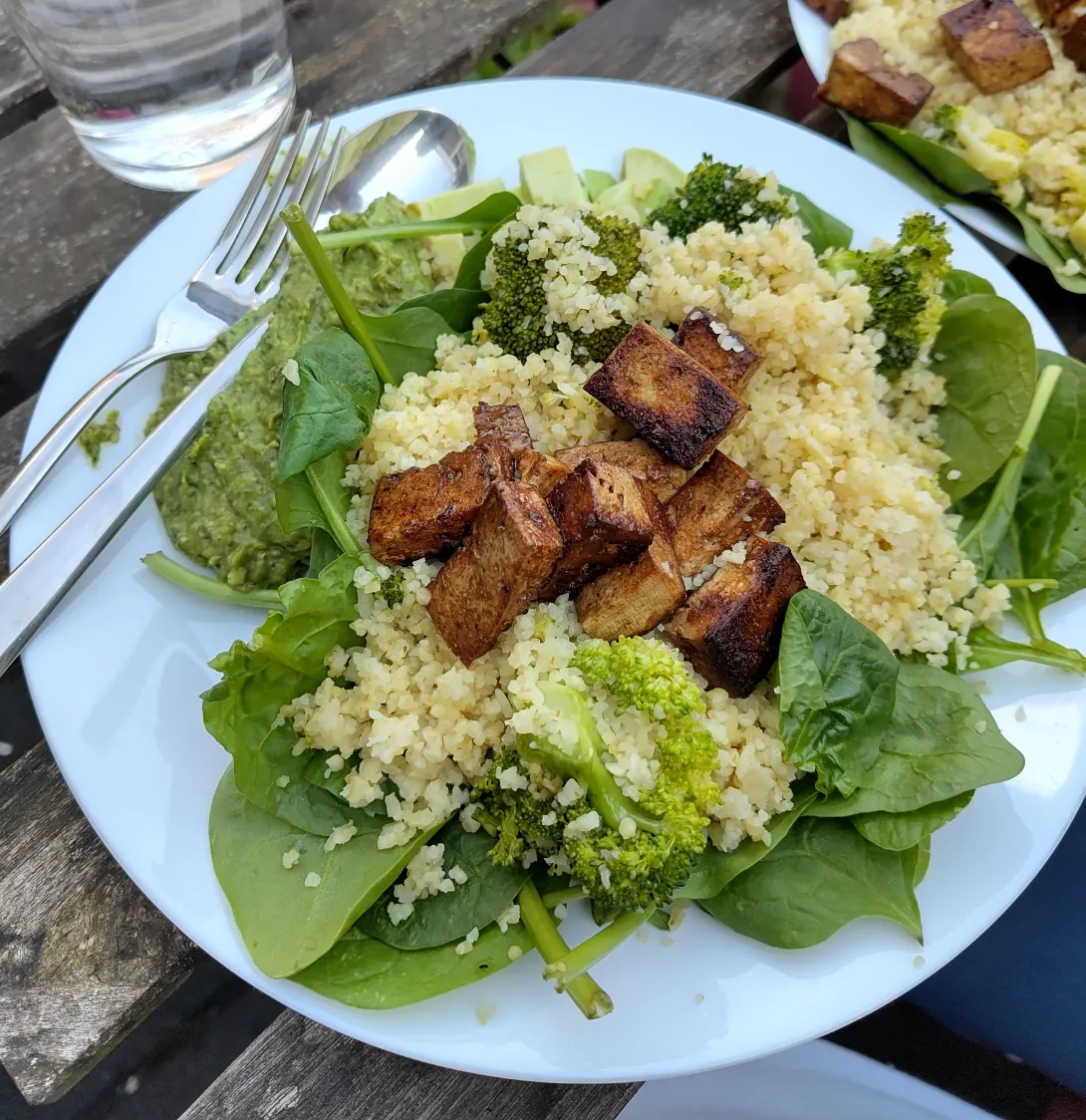 Een bord met bulgur en tofublokjes geserveerd op verse spinaziebladeren met daarnaast de broccoli-pesto.